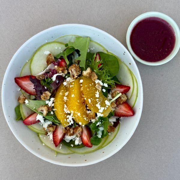 A vibrant salad with mixed greens, orange slices, strawberries, walnuts, feta, and a side of dressing.