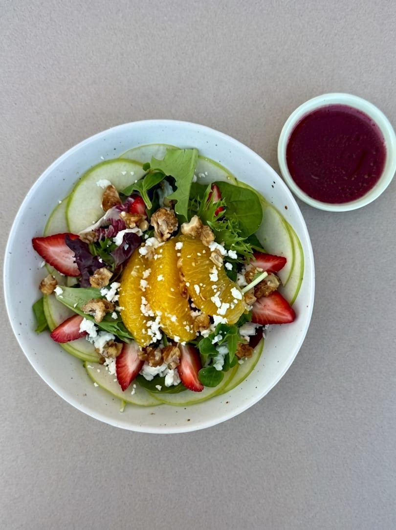 A vibrant salad with mixed greens, orange slices, strawberries, walnuts, feta, and a side of dressing.