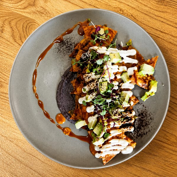 Chilaquiles with red sauce, black bean puree, avocado, crema, and microgreens on a grey plate.