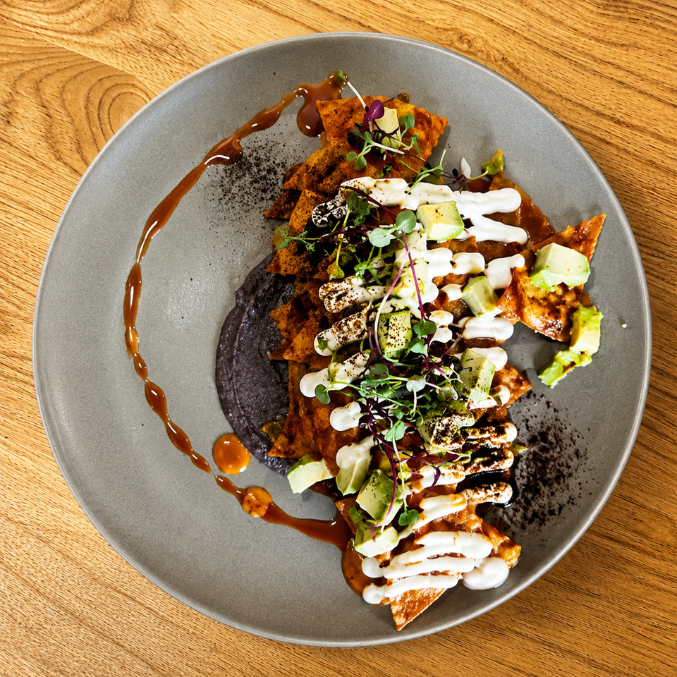 Chilaquiles with red sauce, black bean puree, avocado, crema, and microgreens on a grey plate.